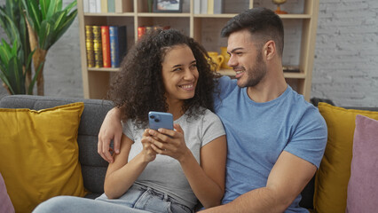 A smiling couple lounges on a sofa using a smartphone in a cozy living room, symbolizing love and connectedness.