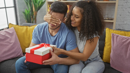 A woman surprises a man with a gift in a cozy living room, symbolizing love and celebration.