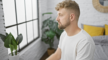 Caucasian man with beard and blue eyes wearing a white shirt indoors, looking pensive in a modern bedroom.