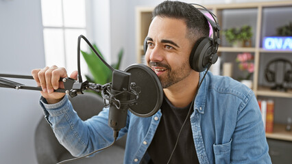 Handsome hispanic man with beard smiling while recording in modern radio studio setup