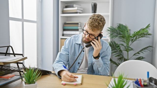 A young caucasian man with blue eyes and a beard makes a phone call while writing notes in a modern office.