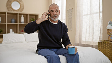A mature man with a beard talks on the phone in a cozy bedroom while holding a mug.