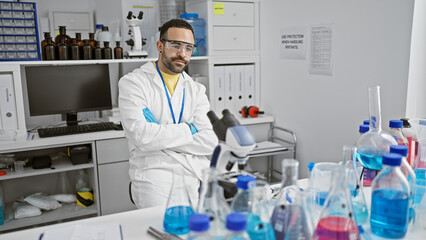 Naklejka premium A bearded hispanic man in a white lab coat stands thoughtfully in a laboratory filled with scientific equipment.