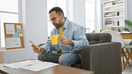 Hispanic man with beard using smartphone and holding cup in modern office setting.