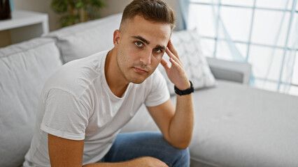 A contemplative young hispanic man sits casually in a well-lit modern living room, exuding a...