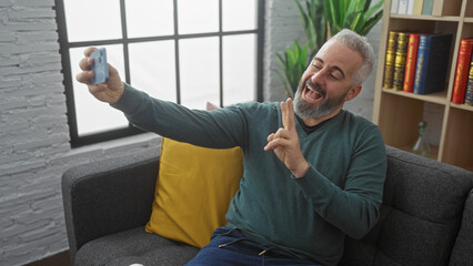 Cheerful bearded man taking selfie with victory sign in cozy living room.