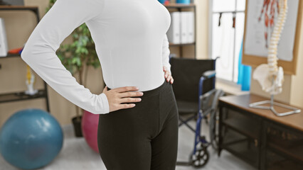 Woman in a physiotherapy room holds her hip, wearing gym attire, surrounded by exercise balls and medical equipment.