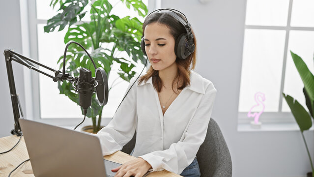 A young hispanic woman wearing headphones speaks into a microphone while working on a laptop in an indoor setting.