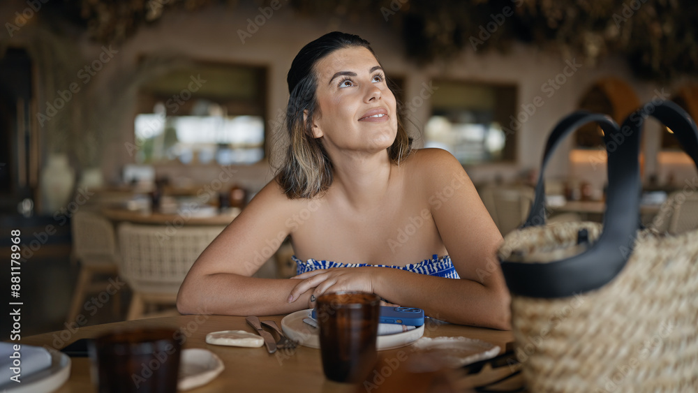 Wall mural Young hispanic woman smiling looking around sitting on the table at the restaurant