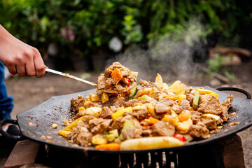 A person stirring a sizzling outdoor stir-fry of beef and mixed vegetables on a large skillet. The dish is steaming hot and ready to serve.