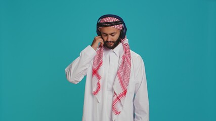 Fototapeta premium Joyful person dancing on songs in studio, listening to modern music on headphones and having fun on camera. Middle eastern adult in traditional clothing showing groovy dance moves.