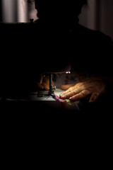 Close-up of an elderly woman’s hands sewing fabric on a machine, illuminated in a dark setting. Concept of sewing.
