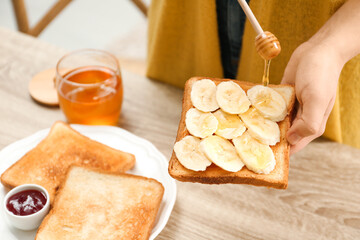 Woman pouring honey onto toast with banana at table indoors, closeup