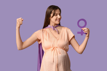 Beautiful young pregnant woman in superhero cape with gender symbol showing muscles on lilac...