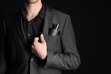 Handsome young man with bottle of perfume in pocket of his jacket on black background. Closeup