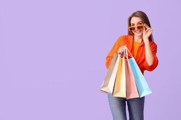Young woman in sunglasses with shopping bags on lilac background