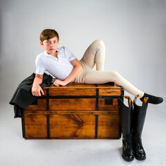 Portrait of a preteen boy equestrian rider casually reclining across the top a long wooden chest or...