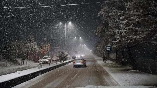 Snow storm traffic on snowy slippery road with cars driving down it and snowflake falling in slow motion. The cars are covered in snow and the street is lit up by street lights.