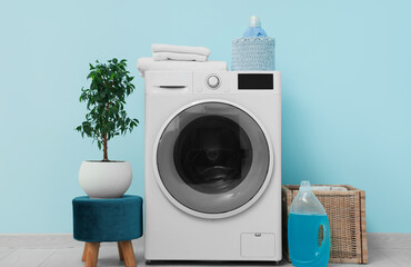 Modern washing machine with houseplant, basket and detergents near blue wall