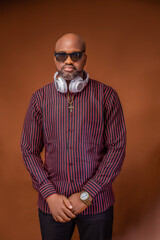 Close up of an African man on a corporate lined shirt and black trouser looking at camera  with headset on neck wearing a wrist watch, gold chain  and sunglasses, brown shoe on a brown background stud