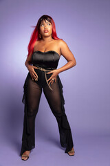 Attractive and stylish black Latina woman with straight red hair wearing low cut black top, see-through flared pants and high heels posing full length on cool purple studio background 