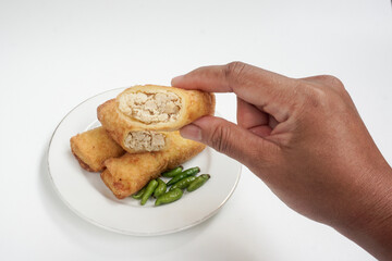 Hand holding ‘Sosis solo’ or fried egg crepe with minced beef or chicken filling. This common Indonesia snack is served with chili pepper on white plate isolated in white background