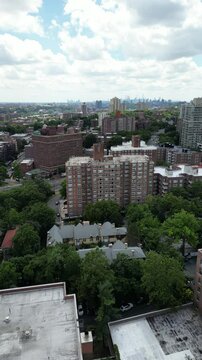 An aerial residential view shows brown apartment buildings among leafy trees, with blue sky and white clouds above the neighborhood. The brick blocks and green trees form a layered urban neighborhood.