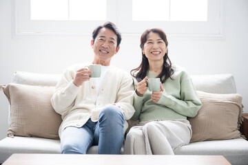 Obraz premium テレビや動画などを見るリビングでくつろぐ50代の夫婦の全身 話す会話するカップルの正面の全身