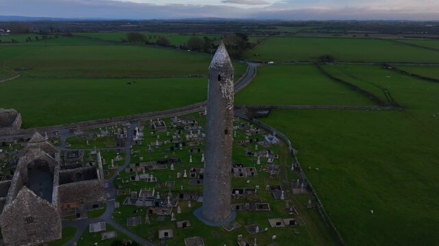 Kilmacduagh Monastery, County Galway, Ireland, November 2023. Drone slowly flies left to right around historic Kilmacduagh Monastery tower, with fields and sky in background.