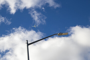 Clear sky backdrop - fluffy white clouds - commercial airplane mid-flight - street lamp foreground. Taken in Toronto, Canada.