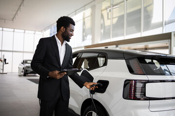 Smiling car dealer in suit demonstrating electric SUV in modern dealership. Concept of clean energy, sustainable transportation, and automotive sales.