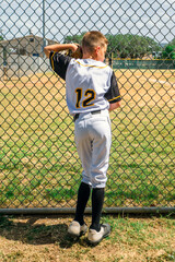 Male youth baseball player in his uniform, standing against a chain link ballpark fence waiting for the game to start