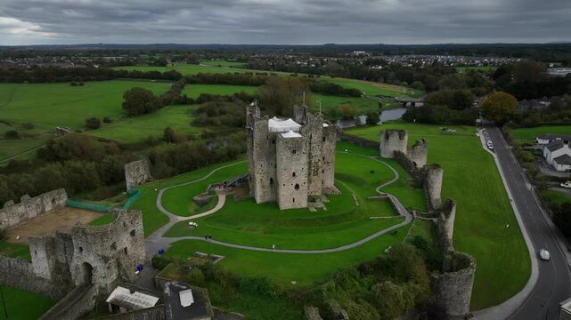 Trim Castle, County Meath, Ireland, October 2023. Drone orbits clockwise and descends towards the Medieval Fortress with greenery ground and road nearby on cloudy day.