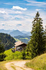 View of an alpine farm and the mountain panorama of the Bavarian Alps (Germany). 
A forest path and a large spruce tree in the foreground. Blue sky with clouds 