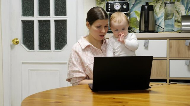 Young woman does not waste time while she sits at home with her child, takes online training or professional development. She sits in kitchen by table in front of her laptop and answers teacher