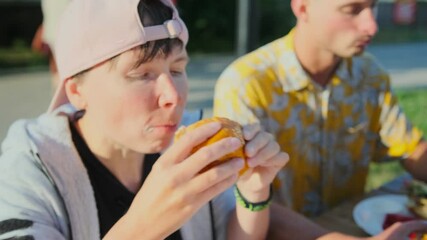 A girl in a cap bites off a burger with appetite while sitting next to a guy in a street cafe with wooden tables. Young people eat fast food in the summer.