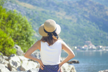 Fototapeta premium Woman tourist during summer vacation in Kotor Bay, Montenegro. A tanned girl standing with her back to the camera looks at the sea coast, holding her hat with both hands - close-up photo.