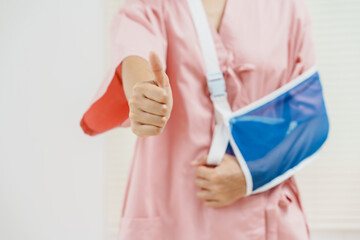 A young Asian female wearing a pink patient uniform stands isolated against a white wall background, showing confidence despite having her arm in a sling due to an injury.