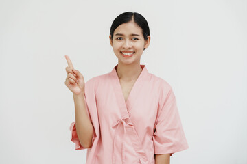 A young Asian female wearing a pink patient uniform stands isolated against a white wall background, showing confidence despite having her arm in a sling due to an injury.