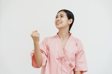 A young Asian female wearing a pink patient uniform stands isolated against a white wall background, showing confidence despite having her arm in a sling due to an injury.