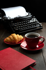 A red cup with coffee. Croissant on a plate. An old vintage typewriter and an empty piece of paper. The notebook is red. The concept of a business breakfast.