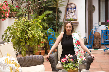 Fototapeta premium Beautiful young woman with brown hair, black suit and silk manila shawl embroidered with flowers, sitting in an armchair in the courtyard of a beautiful hotel. Concept beauty, travel, accommodations.