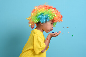Cute little asian boy in clown wig blowing party confetti isolated on blue background. copy space