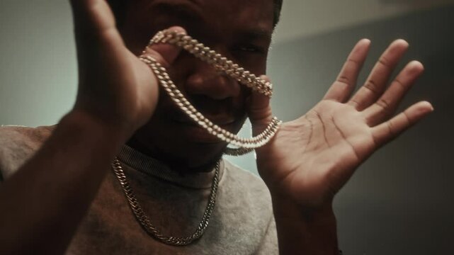 Cool Black guy in baseball cap and set of silver chain necklaces dancing with his arms in projector light on dark background