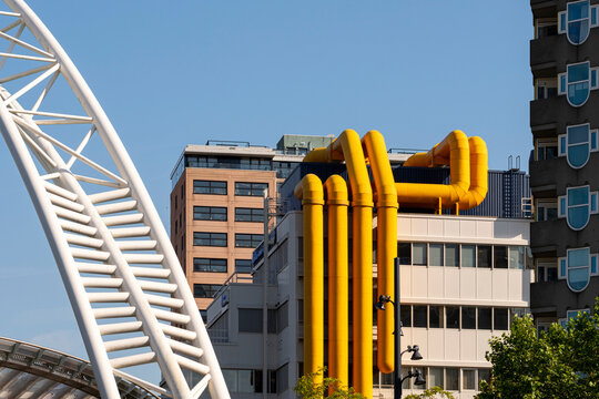 An urban scene featuring modern architecture and industrial elements, including yellow pipes and structures, showcasing a blend of contemporary design and functional infrastructure in Rotterdam