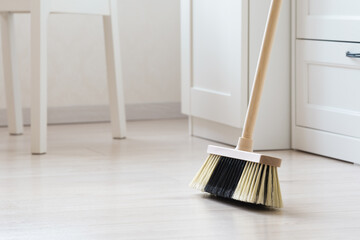 Broom with wooden stick on light laminate floor beside white wooden furniture at home room. Closeup. Cleaning hand tool. Regular cleanup.