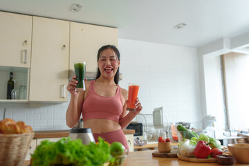 Sporty Asian woman in sportswear holding a glass of vegetable juice. Fruit smoothie after exercise for lively energy in the home kitchen. Morning health concept for athletes, keeping in shape.