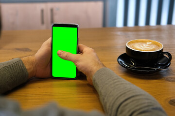 Young man sitting at cafe holding smartphone green mock-up screen in hand. Male person using chroma key mobile phone. Vertical mode. Touching, swiping display, tapping, surfing internet social media
