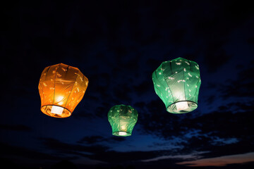 Three sky lanterns in the colors of green, orange and white are floating into the dark blue evening sky