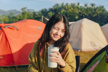 Beautiful Asian woman is sitting outside in a tent with a cup in her hand, winking to the camera.